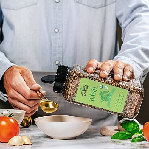 Person pouring organic basil seasoning into a bowl with tomatoes and garlic nearby.